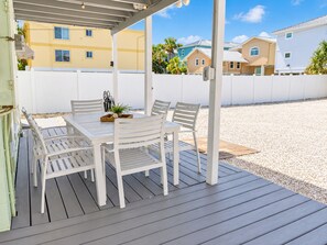 Une terrasse extérieure couverte, avec une table et des chaises, entourée d’une clôture blanche et bordée de bâtiments en arrière-plan.