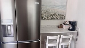 A compact kitchen with a stainless steel refrigerator, a water dispenser, a coffee maker, and two white chairs.