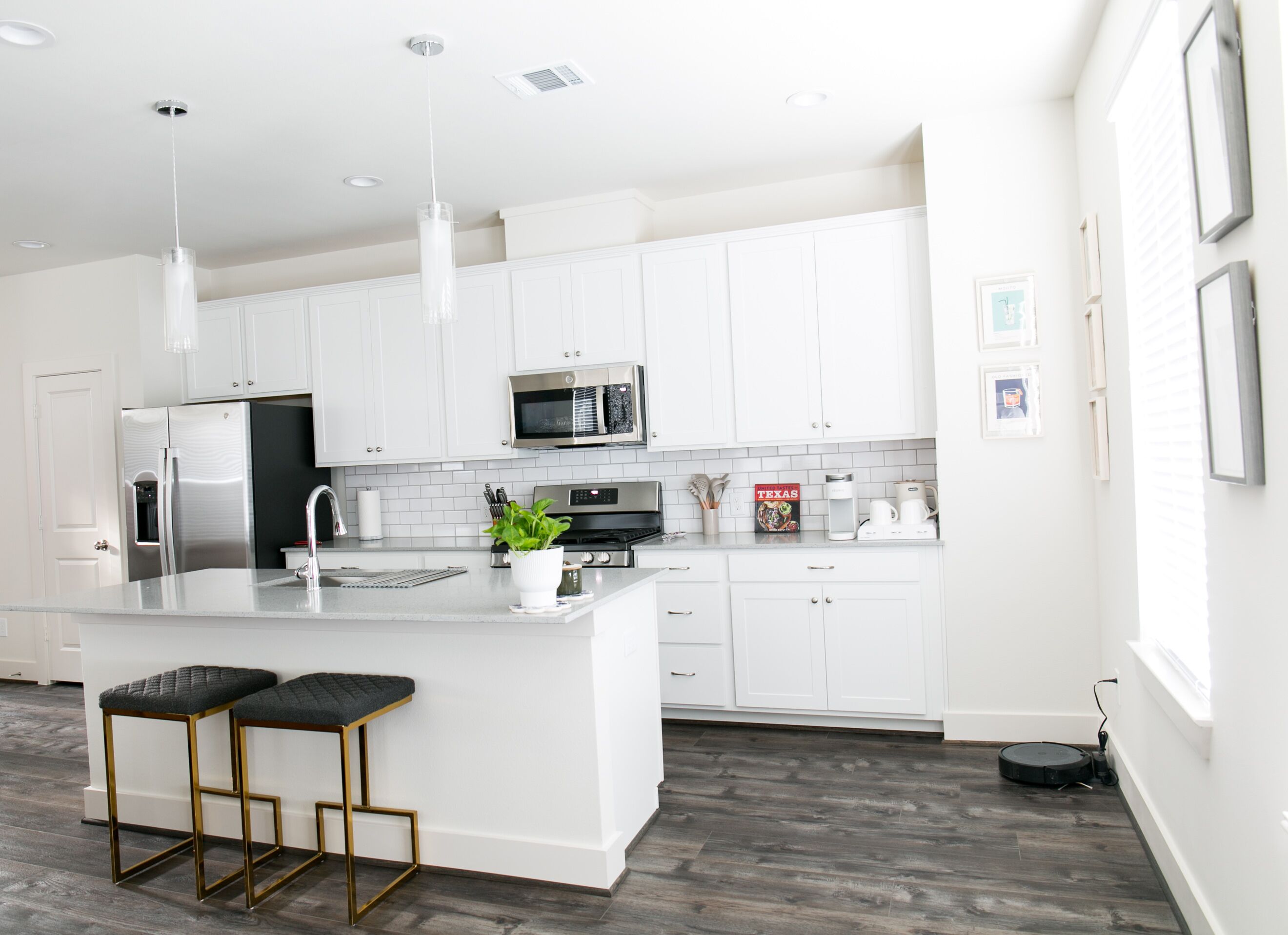 A modern kitchen with white cabinets, stainless steel appliances, a central island with stools, and a robotic vacuum cleaner.