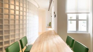 Une salle de conférence avec une longue table, des chaises vertes, un mur en blocs de verre et une fenêtre avec des stores.