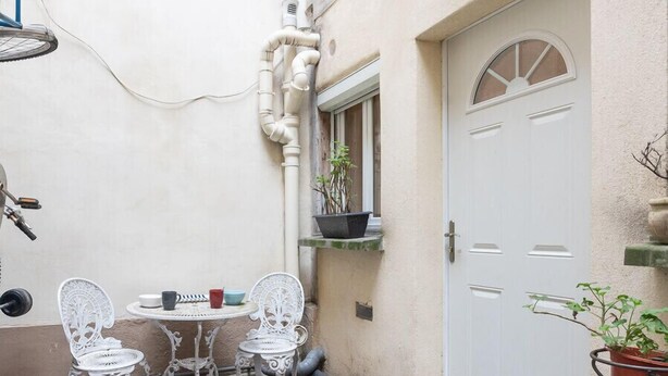 A small outdoor seating area with two white metal chairs and a table, a white door, and a window with a plant on the windowsill.