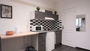 A compact kitchen with a microwave, a sink, and a small refrigerator. There are two potted plants on the countertop, and a trash bin is visible.