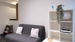 A modern room with a grey sofa, white cushions, a white side table, a glass candle, and a white shelving unit with a plant.