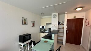 Une cuisine compacte équipée d’un four à micro-ondes, d’une petite table et d’une chaise. Au fond, on aperçoit un meuble blanc avec un évier et une cuisinière.
