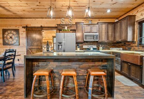 A rustic kitchen with wooden cabinets, a central island with stools, a clock, and a sign that reads 'S&S'.