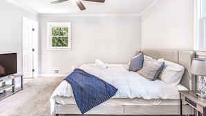 A bedroom with a bed, a ceiling fan, a TV, and a window with a view of greenery.