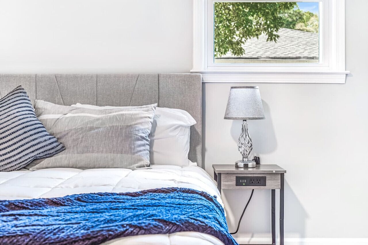 A neatly made bed with a blue blanket, a grey headboard, and striped pillows. There is a bedside table with a lamp and a power outlet. A window with a view of a tree and a building is visible.