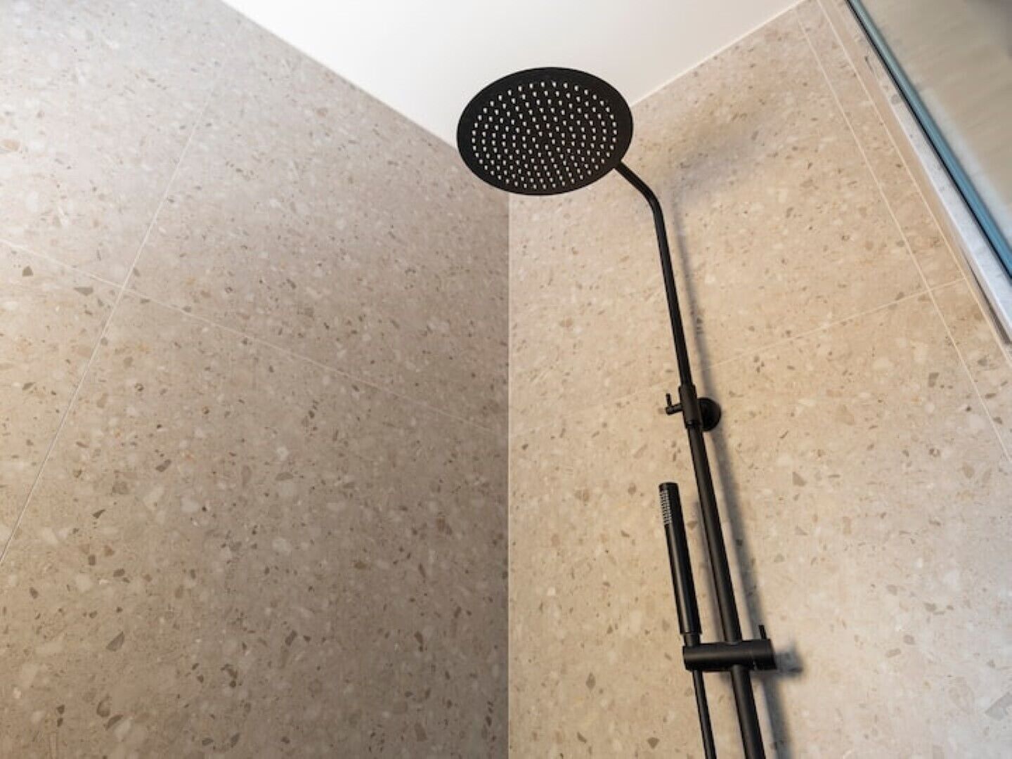 A shower with a round head and a long handle, installed in a room with speckled beige tiles.
