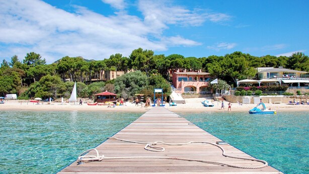 Un molo di legno si protende in acque blu cristalline, con una corda legata, che conduce a una spiaggia con persone, imbarcazioni e edifici sullo sfondo.
