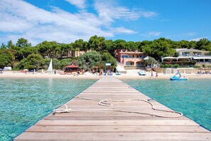 A wooden pier extending into clear blue water with a rope tied to it, leading to a beach with people, boats, and buildings in the background.