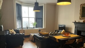 A cozy living room with a black leather sofa, a dining table with chairs, a fireplace, and a large window with blinds. - Brighton Beach Stylish 1 Bed Apartment (Brighton)