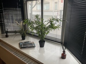 Plants and a book sit on a window sill, offering a view of a nearby building and tree.