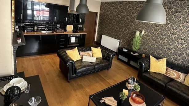 A modern living room with a black leather sofa, a coffee table, and a dining area with a black table and white plates. - Brighton Beach Stylish 1 Bed Apartment (Brighton)