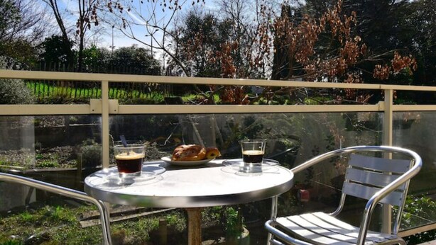 A glass-enclosed outdoor seating area with a table and two chairs, overlooking a garden with trees and a pond.