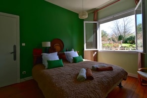Une chambre avec un grand lit, des murs verts, une fenêtre donnant sur des arbres et un plancher en bois.