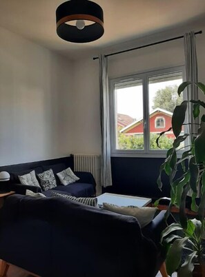 Un salon avec un canapé bleu foncé, une table basse blanche et une grande fenêtre donnant sur une maison rouge.
