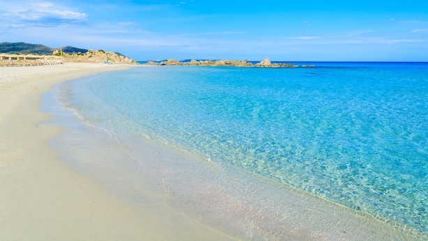 Une plage avec une eau turquoise et limpide, un rivage sablonneux et, au loin, des formations rocheuses.