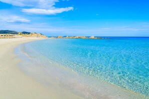 A beach with clear turquoise water, sandy shore, and rocky outcrops in the distance.
