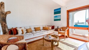 Ein gemütliches Wohnzimmer mit einem weißen Sofa, Korbstühlen, einem Couchtisch und einem großen Fenster mit Blick.