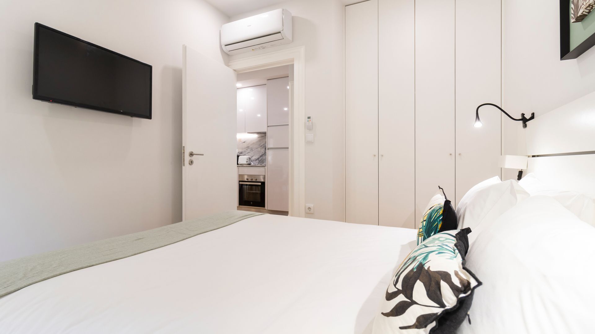 A bedroom with a bed, a TV mounted on the wall, and a kitchen visible through an open door.