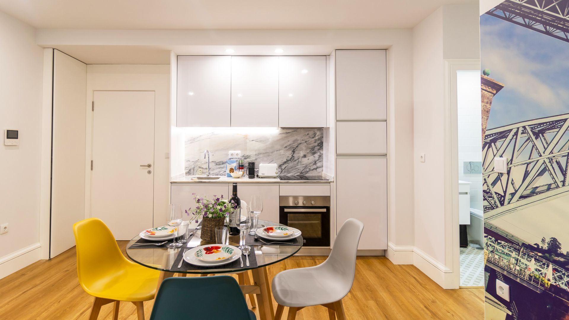 A compact kitchen with white cabinets, a marble backsplash, and a dining area with a glass table and colorful chairs.