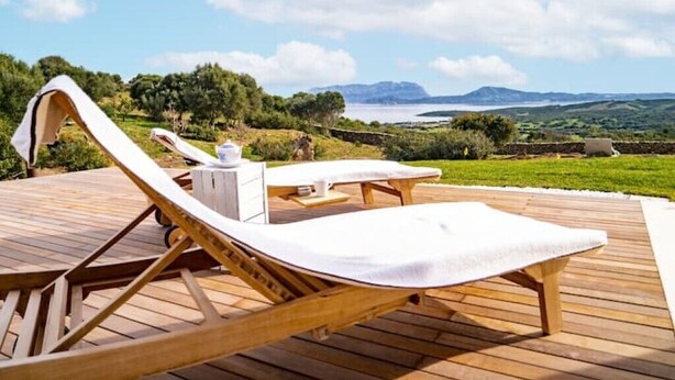 A wooden deck with lounge chairs and a table, overlooking a scenic landscape with hills and a body of water. - Pittulongu Olbia Residence A Domo Mea Retreat (Pittulongu)