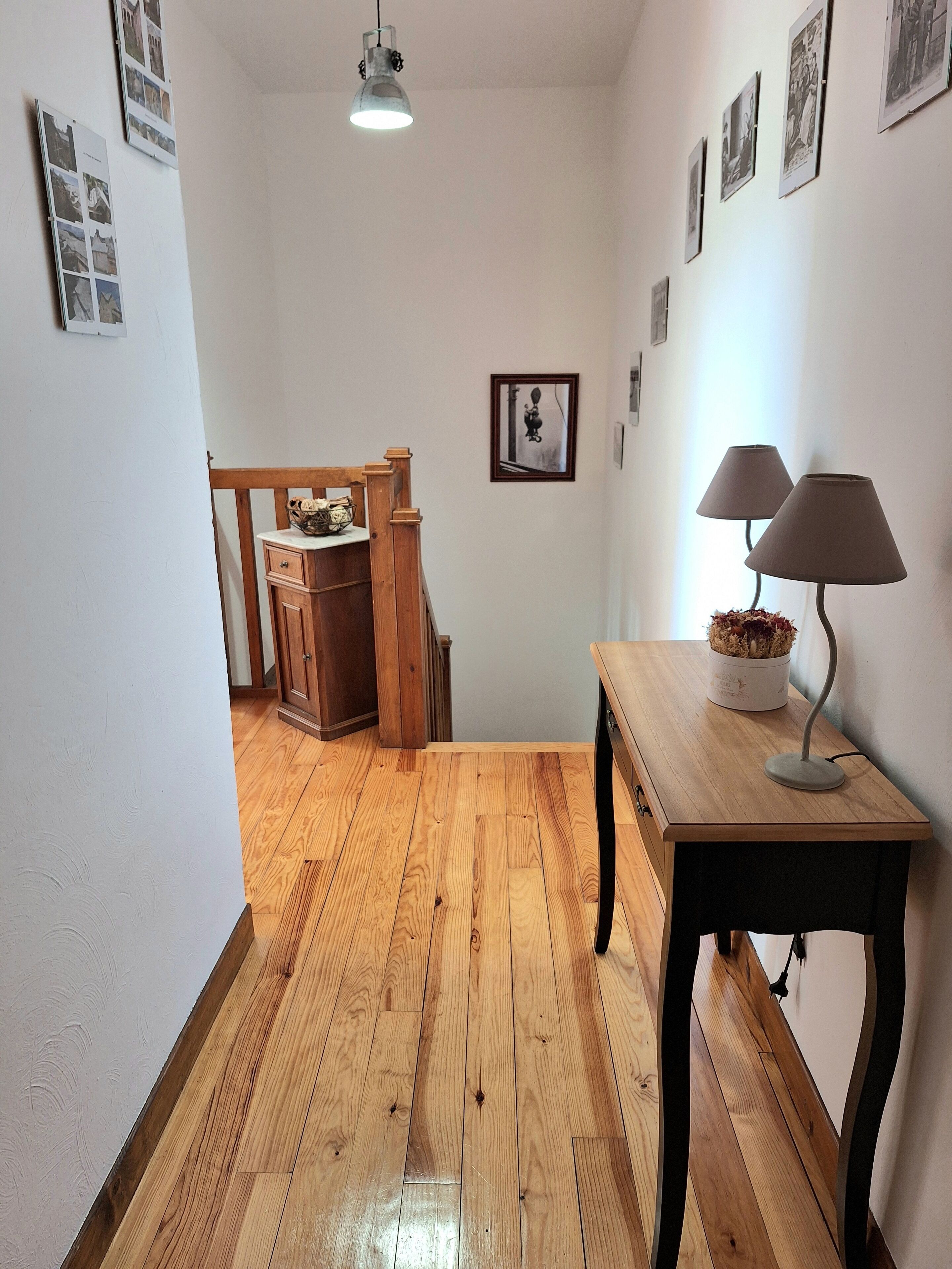 A hallway with wooden flooring, a wooden chair, a small table with a lamp, and framed pictures on the walls.
