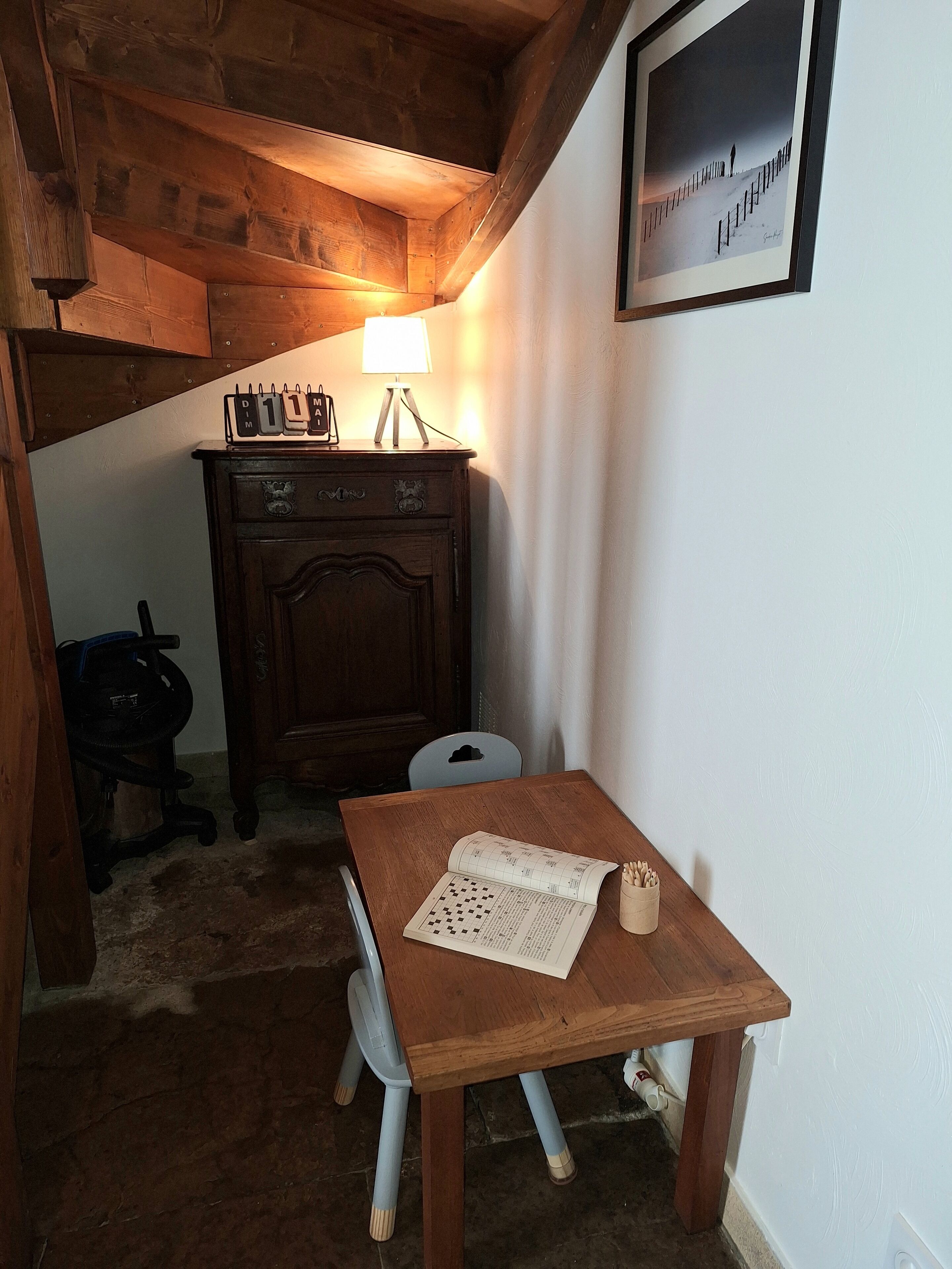 Una mesa de madera con un calendario, una lámpara, una silla, una lámpara de escritorio y un cuadro en la pared.