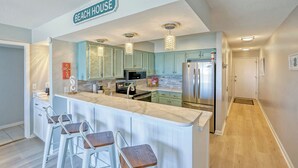 Een keuken met een marmeren eiland, barstoelen, turquoise kastjes, roestvrijstalen apparatuur en een bord met 'BEACH HOUSE'.