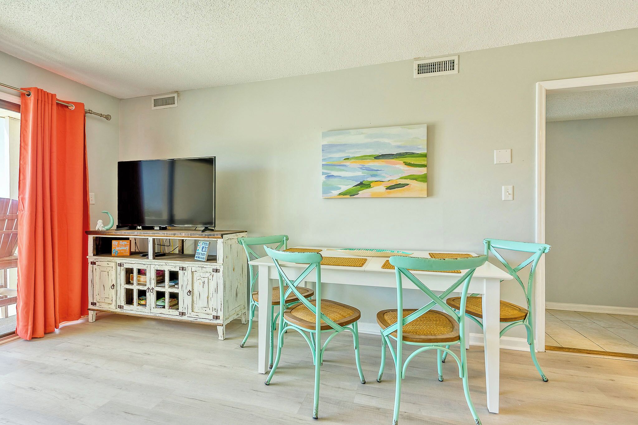 Uma sala de jantar com uma mesa branca, quatro cadeiras turquesa, uma televisão sobre um armário branco e um quadro na parede.