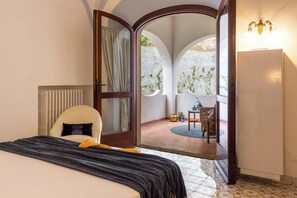 A bedroom with a bed, a chair, a door leading to an outdoor patio with a table and chairs, and a refrigerator.