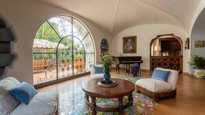 Un salon spacieux avec un piano, une table ronde en bois et une grande fenêtre en arc d’où l’on peut admirer un jardin.