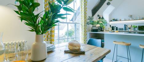 Un comedor con una mesa de madera, sillas, un florero con una planta, hogaza de pan y una ventana con cortinas amarillas.