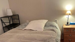 Una cama bien hecha con una colcha gris, una mesita de noche con una lámpara, y una lámpara de pared.
