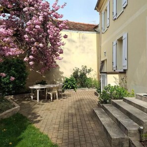 Une cour avec un chemin en briques, une table et des chaises, et un arbre en fleurs.