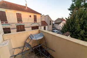 Un balcon avec une chaise en métal et une table, donnant sur une rue bordée d’immeubles et d’un arbre.