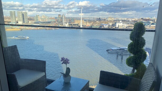 A balcony with wicker chairs and a small table with a flower vase, overlooking a river with boats and a cityscape in the background. - Stunning 1-bed Apartment in London With River View (London)