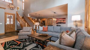 A spacious living area with a gray sofa, a wooden coffee table, and a patterned rug. A staircase with wooden railings leads to an upper level. - Old Greenwood Townhome 13C (Truckee)