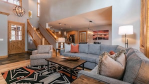 Un vaste espace de vie comprenant un canapé gris, une table basse en bois et un tapis à motifs. Un escalier avec des rampes en bois mène à l’étage supérieur.