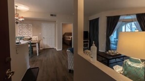 Sala de estar con sofá, mesa de centro y lámpara. Hay una puerta que da a una zona de cocina con mesa y sillas.