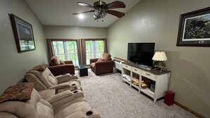 Ein Wohnzimmer mit einem beigefarbenen Ecksofa, einem Fernseher auf einer weißen Kommode und einem Deckenventilator.