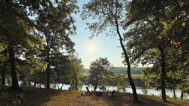 Ein Campingplatz am Seeufer mit Picknicktischen und -stühlen, umgeben von Bäumen und blauem Himmel.