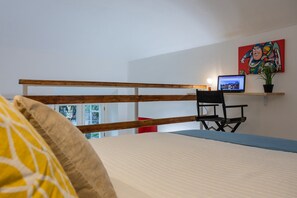 A bedroom with a bed, a desk with a laptop, a chair, a red box, and a colorful painting on the wall.