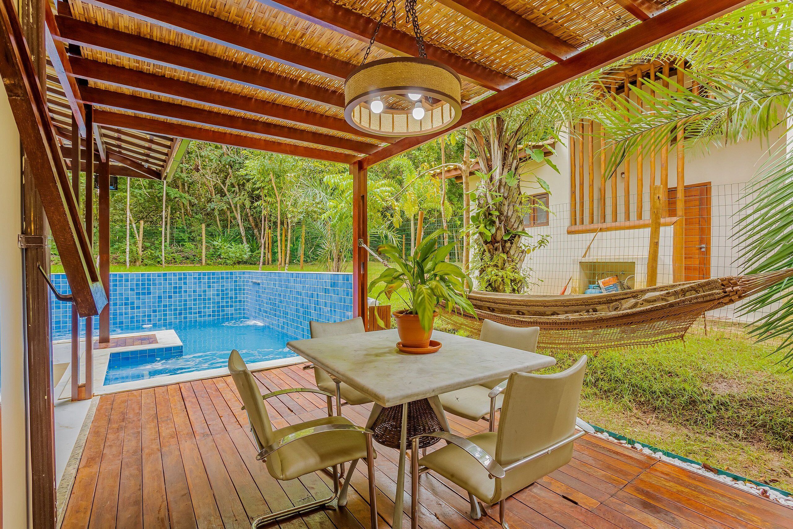 A wooden deck with a table and chairs, a hammock, a pool, and lush greenery.