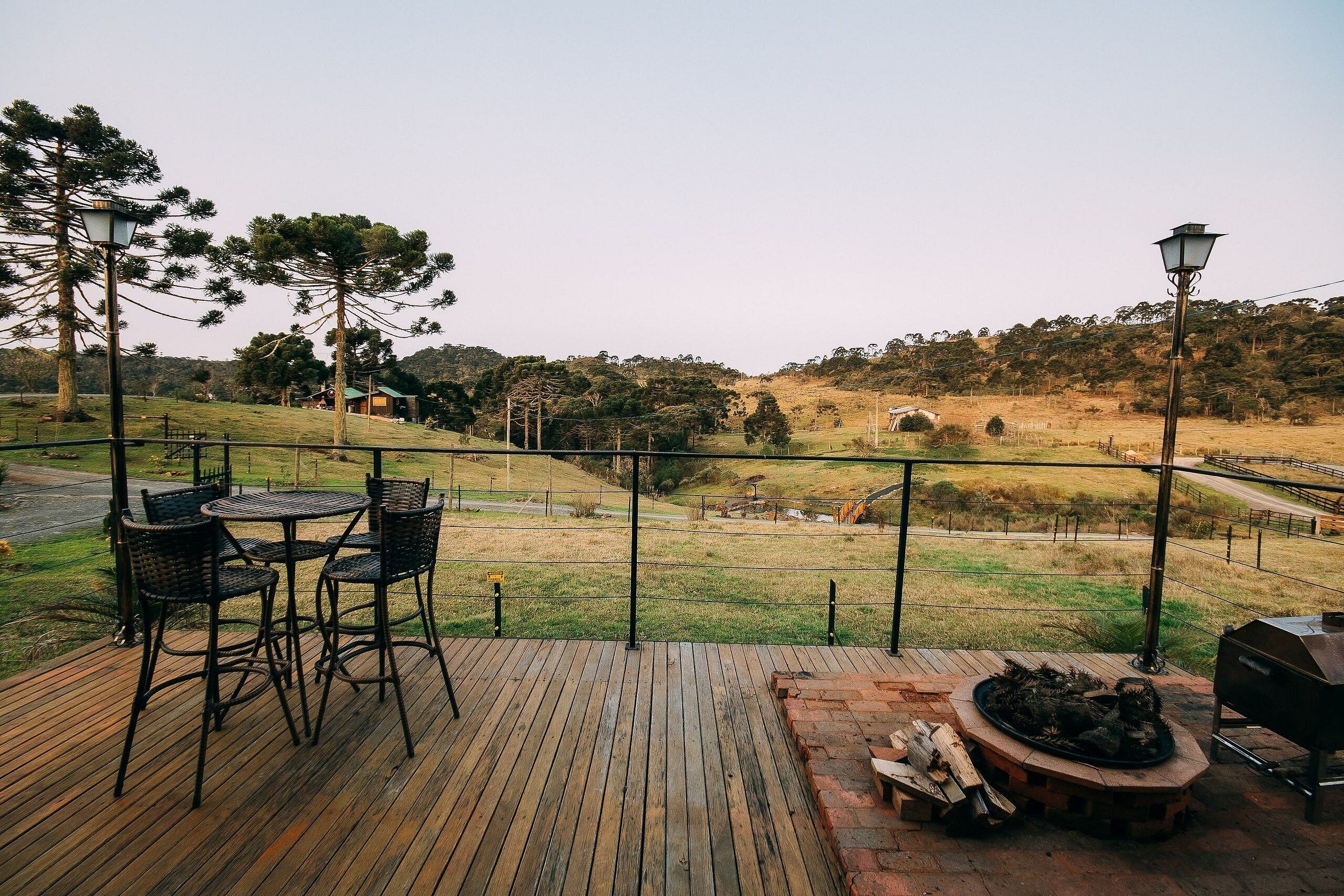 Terraza de madera con mobiliario exterior y zona de barbacoa, con vistas a un paisaje cubierto de césped, árboles y una colina distante.