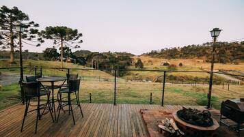 Terraza de madera con mobiliario exterior y zona de barbacoa, con vistas a un paisaje cubierto de césped, árboles y una colina distante.