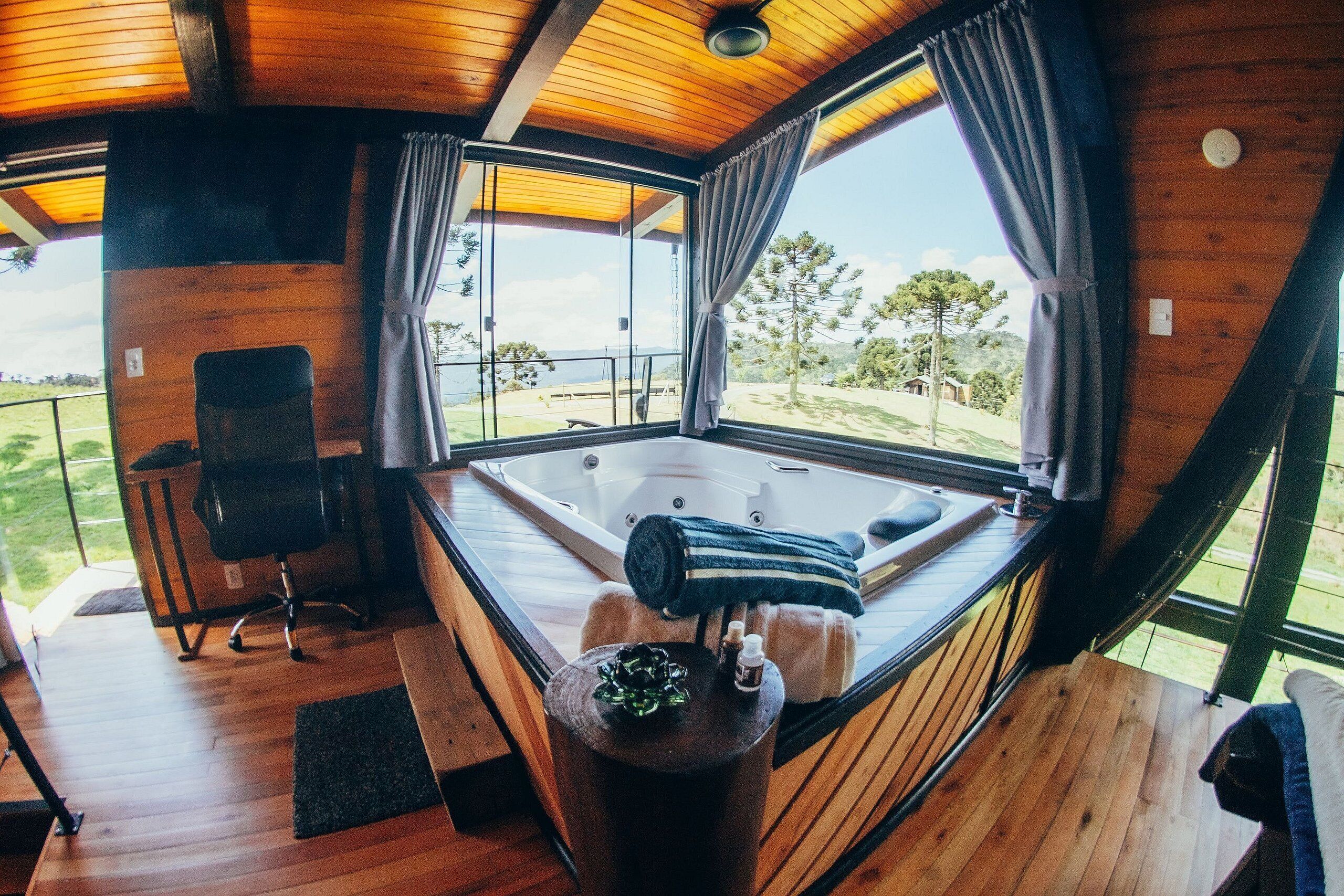 A wooden cabin with a large window, a hot tub, a desk, and a chair.