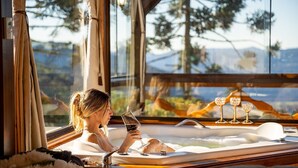 A woman relaxing in a hot tub with a glass of wine, large windows, and a view of trees and mountains. - Sítio Mar de Nuvens Chalés de Luxo  (Urubici)