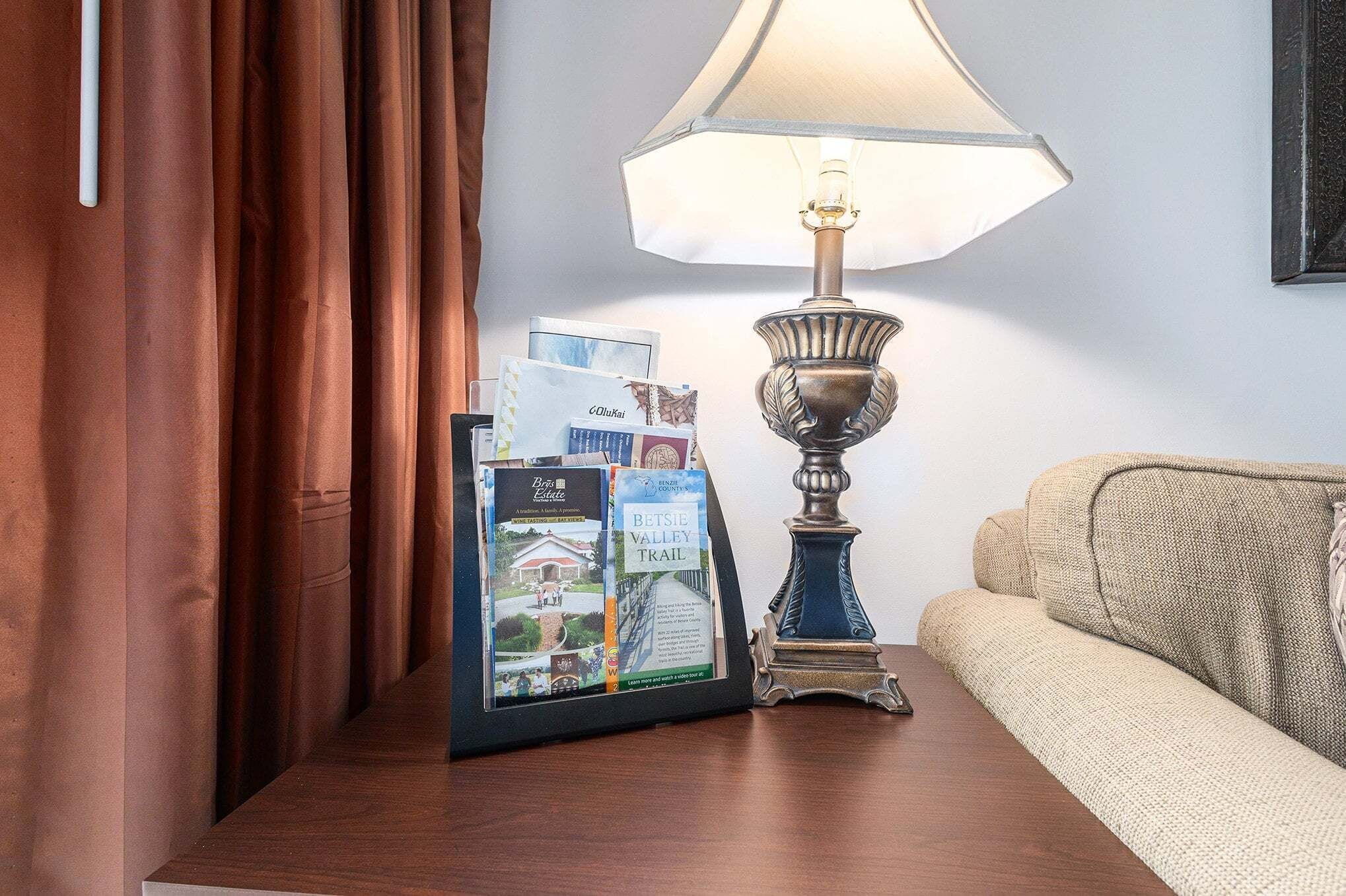A framed brochure stand with travel guides, a table lamp, and a sofa in a room with curtains.