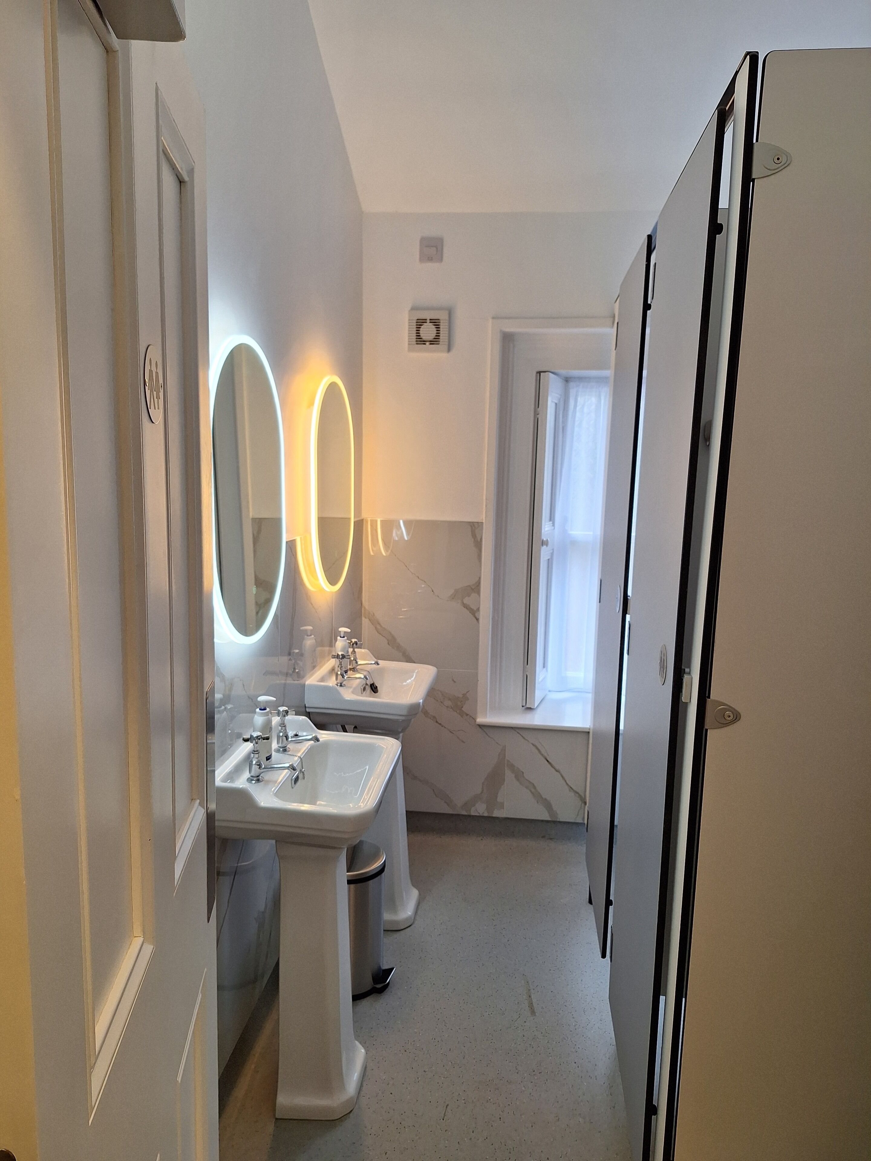A modern bathroom with two sinks, a mirror with illuminated lights, and a door leading to another room.